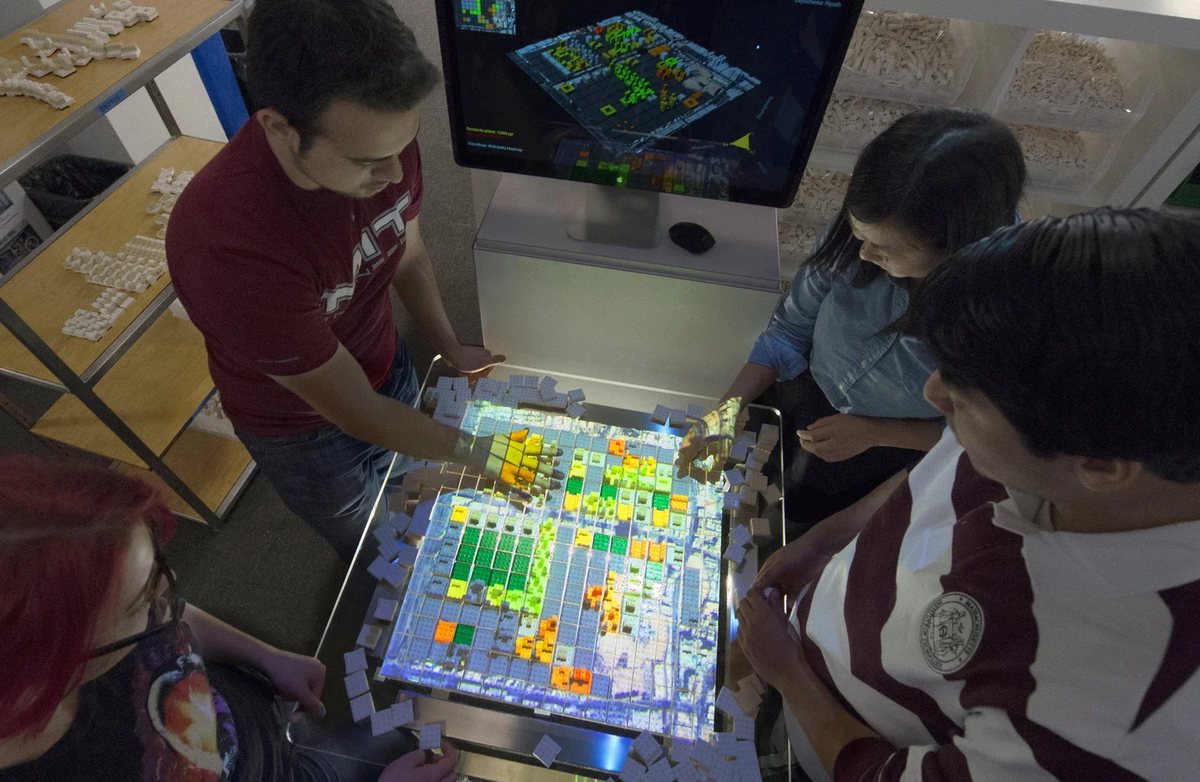 Four people gather around a Tactile Matrix table, rearranging projected Lego blocks of a city model
